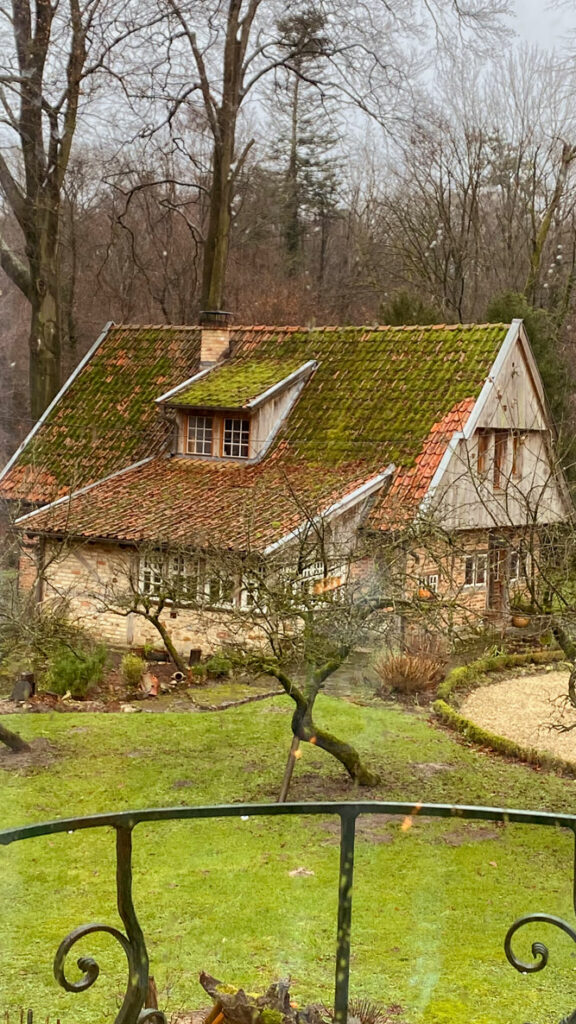 Originalfoto der Hausansicht. Altes Jägerhaus mit spitzem Dach und Anbau. Moosbewachsen und mit Stein gemauert steht es an einer grünen Wiese mitten im Wald.